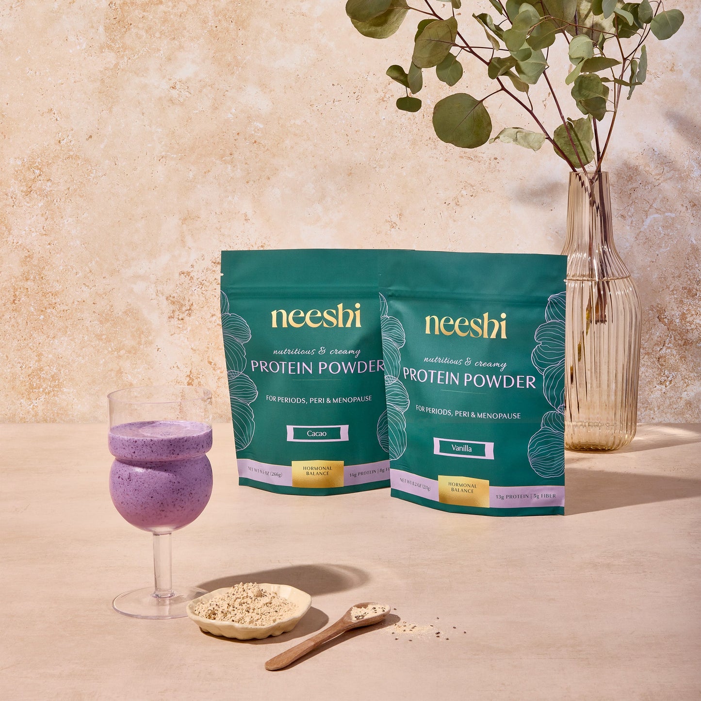 Two packages of Neeshi protein powder beside a glass of protein shake and a small bowl of oats, with a plant in the background.