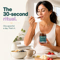 A woman in a white ribbed tank top holds a jar of Neesta Organic Spinach Spread and eats from a spoon while standing in a bright kitchen. Text on the left reads, 'The 30-second ritual.