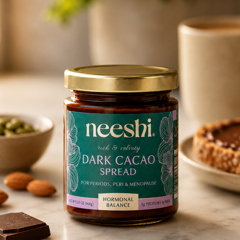 Jar of neeshi dark cacao spread on a table with a cup, bowl, and chocolate pieces.